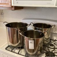 Three stainless steel stock pots of different sizes sitting on a gas stove burner. Each pot has side handles and a matching lid with a handle. Reflective surface with no visible brand markings.
