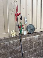 Metal red/green bird yard ornament on black stake with small bell hanging below, placed leaning against a beige fence over a brick wall.