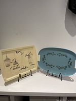 Two serving trays displayed on stands against a white background, one cream-colored with black birdcages and birds, the other blue with a leaf pattern.