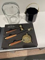 Set of four gold tone and wood handled bar tools arranged in a black foam tray, accompanied by two vintage ice buckets: one clear heavy crystal with silver handle, one black glass with gold handle.