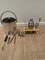 Photo showing entire lot with ice bucket on the left, multiple metal jiggers, muddler, corkscrew, tongs and shaker set with accessories on wooden stand on the right.