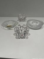 Five clear glass candle holders displayed on a white surface. Two have Kosta Boda floral petal styles. One has a circular textured design. One tiered dish, and one rounded floral shape candle holder.
