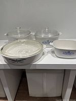 Photo showing one clear glass Anchor Hocking Wheat serving bowl with lid and four Corningware Cornflower Blue white ceramic casserole dishes with lids or without lid in a group display.