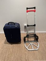Photo of blue fabric wheelie suitcase with handle, standing next to folded hand dolly with red and black handles and metal frame on floor.