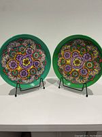 Two round green tin trays with colorful 70's style floral designs on black metal stands shown side by side.