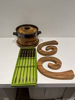 Photo of vintage Laurentian bean pot with lid placed on one wooden spiral-shaped trivet, two pieces comprising the trivet set, and the set of six fondue forks in green box.