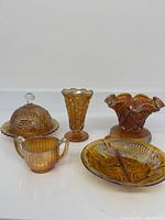 Photo of the entire collection showing a covered butter dish, footed vase, ruffled bowl, creamer, and divided relish dish in marigold carnival glass.