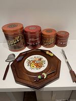 Photo showing four tin canisters in varying sizes with labels Fluffy, Toast, Coffee, and Sugar, accompanied by an octagonal wooden cheese tray with ceramic tile, four fondue forks, and cheese utensils.