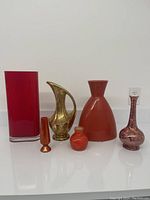 Six vases arranged in a row showing variety in shape and color including red glass, gold ceramic pitcher, and orange ceramic vases.