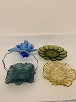 Top-down view of four assorted coloured glass items including a blue wave-design bowl, green bubble-petal dish, square textured green bowl with handle, and amber ruffled dish.