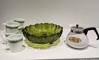 Five white mugs with olive green floral pattern, one Corningware teapot with floral design, and avocado green footed fruit bowl arranged on white surface