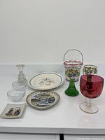 Assorted kitchen and bar items shown together including glass bowls, decorative plates, goblet, decanter, candleholder, and wire basket