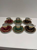 Top-down view of 6 demi tassel cups and saucers arranged in two rows showing vibrant colors and gold markings on each cup and saucer.