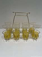 Front view of the set of eight glasses in the gold-tone wire caddy, showing clear glasses with white Greek key patterns and yellow bands around the bottom, all seated in the caddy with handle.