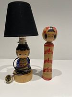 Photo showing entire lot - the black lampshade Kokeshi doll lamp with plug clothed painted Kokeshi figure base, and the traditional signed wooden Kokeshi doll with red and black floral painting.