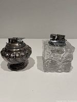 Two vintage tabletop lighters side by side on a flat surface: left is silver plated Ronson Vera-flame Crown lighter with ribbed design, right is clear textured Murano Crystal lighter with chrome mechanism.