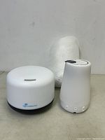 Two white aromatherapy diffusers side by side on a white surface, one short and round, one tall and slim.