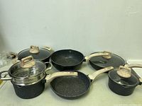 Full view of 7-piece cookware set including pots, pans, lids, and steamer insert. Displays speckled black coating and wood-effect handles.