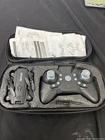 Compact foldable drone and black remote controller in a black foam-lined zippered carrying case with instruction manual visible in mesh pocket.