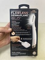 Photo of the front of the Finishing Touch Flawless Dermaplane Glo packaging showing the device image, branding, and key features including LED light, 6 replacement heads, and AAA battery power.