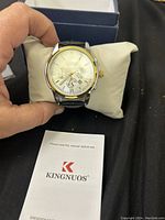 Close-up of KINGNUOS watch face showing silver dial, gold and silver case, date window, and sub-dials with user manual in front.