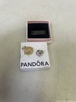 Two Pandora-style charms displayed on a damaged white and pink Pandora storage box, showing charm details and box condition.