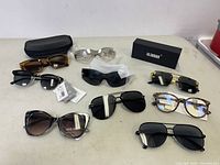 Lot of 8 sunglasses displayed on table with some still in plastic or packaging and one black protective case