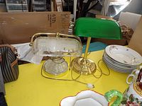 Photo showing both desk lamps on a yellow surface: one with a green glass shade and brass base, the other with a frosted pressed glass shade and matching glass base.