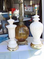 Three vintage lamps displayed on table: two white porcelain lamps with floral embossing and gold-tone bases, and one amber glass lamp with brass base.