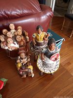 Photo showing all four red clay figurines arranged on the floor in front of a red leather chair. One piece has multiple smaller figures together, the other three are individual female figurines in traditional clothing with accessories.