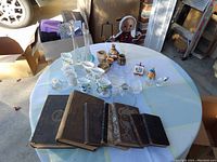 Overview of the table showing antique books, small pottery pieces, vases, glassware, and small collectibles arranged on a round table with white and pastel tablecloth.
