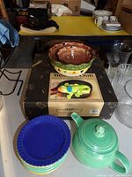 Set of colorful Temptations stacking bowls on a box with a lizard-shaped salsa & chip bowl set placed on top, Fiesta blue rimmed plates stacked and green teapot seen in front