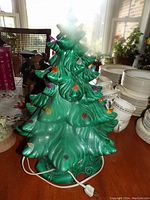 Green ceramic Christmas tree lamp with colorful bulbs shown alongside white china dishes on a wooden table.