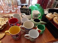 Overview of eight assorted novelty coffee mugs with various designs and colors, positioned on a table alongside other household items.