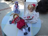 Set of three dolls on light blue and green tablecloth: porcelain doll in white dress holding basket, smaller doll in blue dress with bonnet, and marionette puppet lying down.