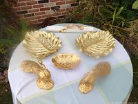Two large golden weeping palm leaf plates, two smaller decorative golden items laid on a white tablecloth with a brick wall background.