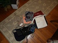 Six handbags arranged on a beige rug with hardwood floor showing, highlighting different colors and materials, including white leather, black leather, black patent leather, navy blue, red cloth, and patterned strap cloth handbag.