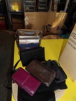 Photo showing seven handbags and wallets arranged on a round basket and yellow box, including black woven handbag, brown wallet, magenta wallet, and smaller black pouch.
