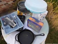 Overview of kitchen lot items on round table in outdoor setting including divided serving dish, utensil set in plastic container, cake pans, muffin tray, lunchbox, and cast iron skillet