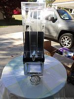 Photo of clear acrylic rectangular beverage dispenser with metal stand and black top, seen outdoors on a table with vehicle in background.