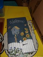 Blue slate welcome sign with birdhouse, sunflowers, white picket fence, and rope hanger