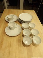 Set showing six white ceramic snack plates with gold rims stacked and six matching white cups grouped together on a wooden table.