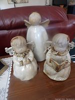 Three ceramic angel figurines on a wooden table in front of a red leather couch.