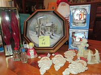 Photo showing large framed winter scene plate, red candles, various Santa ornaments and figurines, unopened night light boxes, and glass candle holders.