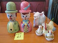 Photo showing the set of colorful vintage Mom and Pop ceramic egg cups (salt and pepper shakers), bird ceramic salt and pepper set with ladle, and ceramic cow creamer on wooden table with dark wood cabinet background.