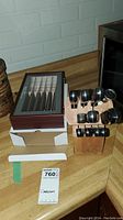 Photo showing a wooden chef's block holding multiple metal utensils with black handles beside a wooden box containing carving knives and a knife sharpener on a kitchen countertop.