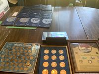 Full view of all coin collections on a wooden table including US 50 State bronze coin set in board, Franklin Mint Zodiac books, Canadian War Collection framed case, and another Canadian collection.