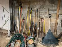 Wide view of the assortment of garden tools against a basement wall showing shovels, rakes, hoes, pruning clippers, and manual jacks