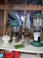 Shelf with four lanterns: white ceramic lantern base, metal oil lamp with green top, amber base oil lamp with glass chimney, and green Coleman propane lantern.