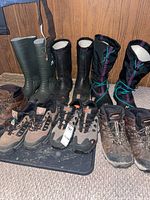 Photo showing seven pairs of men's footwear including winter boots, insulated rubber boots, work boots and work shoes all size 10 clustered on the floor against a panel wall.